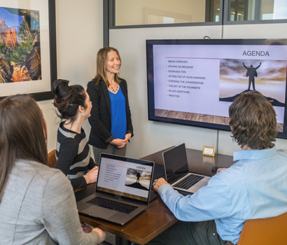 A businesswoman discussing the agenda shown on a flat-screen monitor to three other people inside a meeting room